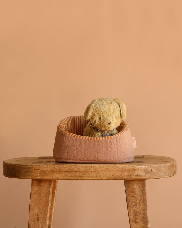 The Maileg Puppy in Basket - Soft Sand features a vintage dog plush in a small, round, sand-colored basket placed on a wooden stool with a plain beige background.