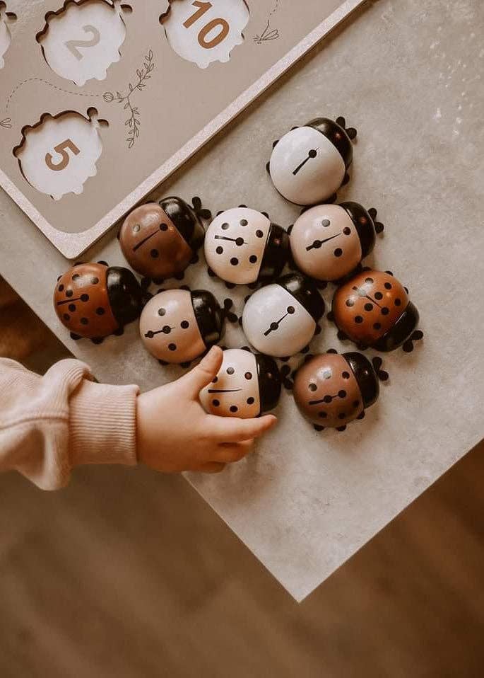 A child’s hand reaches for the My Little Lady Bug Wooden Puzzle, featuring ladybug-shaped wooden pieces in various shades, arranged on a table alongside a Montessori counting toy and a wooden peg puzzle with numbers and animal shapes.