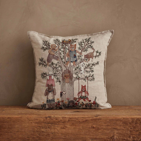 The Coral & Tusk Tree of Fun Pillow features a cheerful embroidered design of a fox, bear, and rabbit playing around a tree on beige fabric, displayed on a wooden surface.