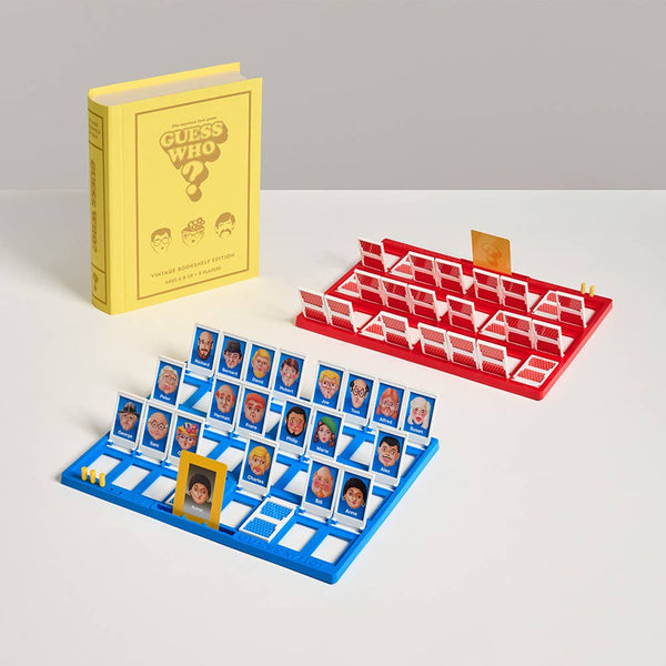 Two Guess Who? Vintage Bookshelf Edition game boards, one red and one blue, are set up on a white surface. A yellow box displaying the product name stands upright in the background, emphasizing its classic design and nostalgic feel.