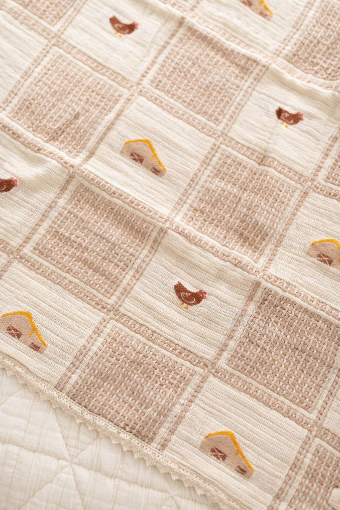 The Organic Cotton Baby Blanket - Farm features a beige woven checkered pattern with small embroidered chickens and yellow-roofed houses. Its textured fabric is finished with charming scalloped edges for an adorable touch.