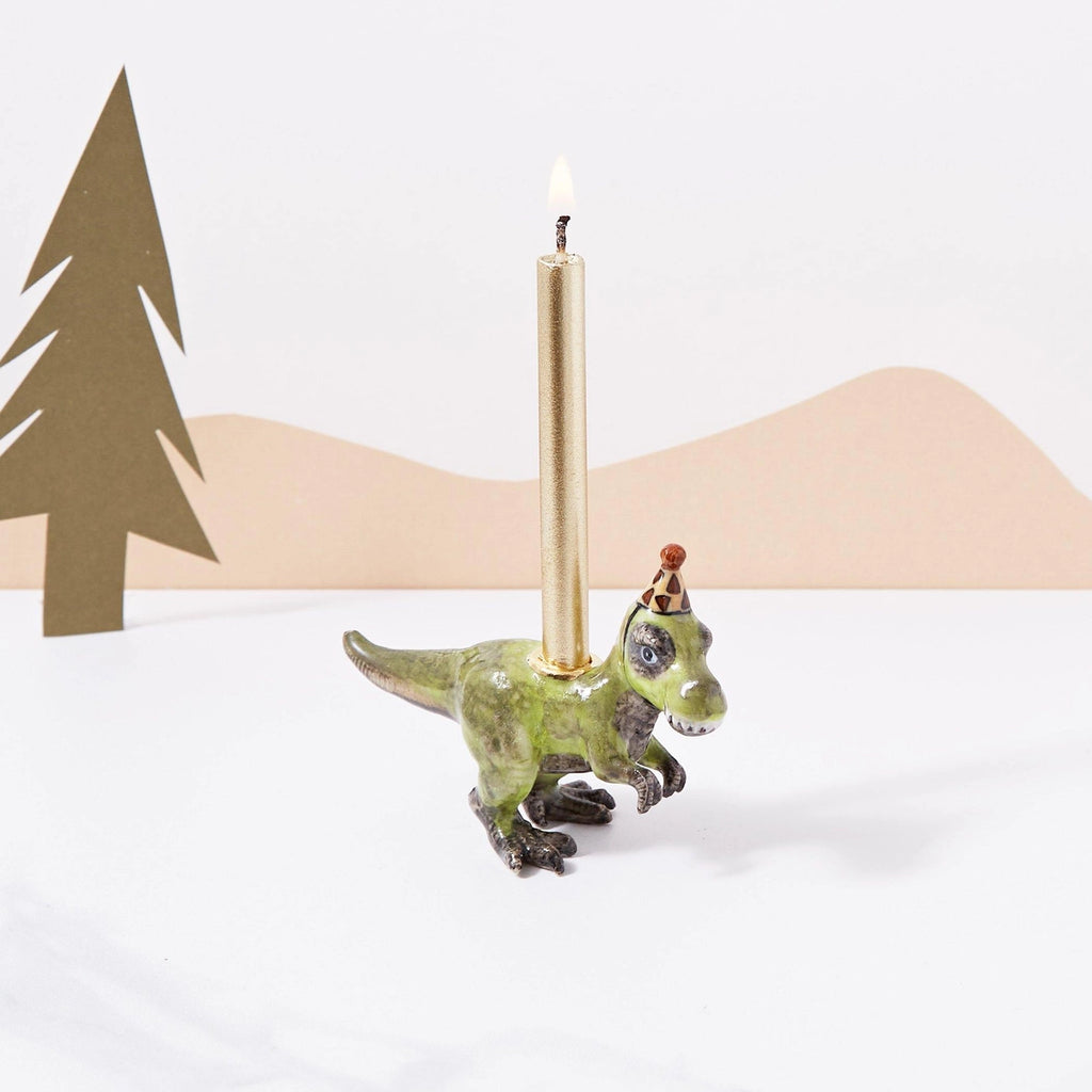 A whimsical T-Rex "Party Animal" Cake Topper shaped like a green dinosaur with a golden taper candle on its back, set against a simple backdrop with paper cut-outs of trees. This porcelain keepsake is hand-painted.