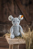 The Steiff Cheesy Mouse Stuffed Animal sits on a straw-covered wooden bench with a rustic wood backdrop and tall wheat-like grass in the foreground.