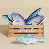 The Market Fish Wooden Play Market Crate features a small wooden crate labeled "Le Toy Van" filled with blue-toned toy fish for pretend play, with one toy fish placed in front of the crate.