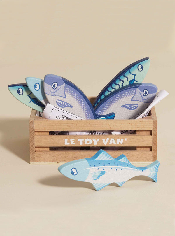 The Market Fish Wooden Play Market Crate contains four blue and white pretend fish and a rolled-up paper, with one fish placed in front of the crate on a light background.
