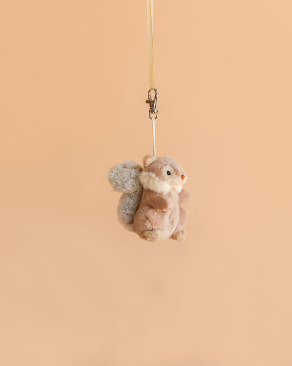A Bukowski - Wiggles Squirrel Bag Charm dangles from a beige ribbon against a plain peach-colored background, making it the perfect woodland accessory for your bag.