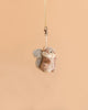 A Bukowski - Wiggles Squirrel Bag Charm dangles from a beige ribbon against a plain peach-colored background, making it the perfect woodland accessory for your bag.