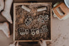 Children’s hands and feet surround a box filled with dirt and wooden construction vehicle shapes, along with toy trucks nearby—creating the perfect setup for engaging sensory play with our Construction Cutter Set - Eco Playdough Tool.