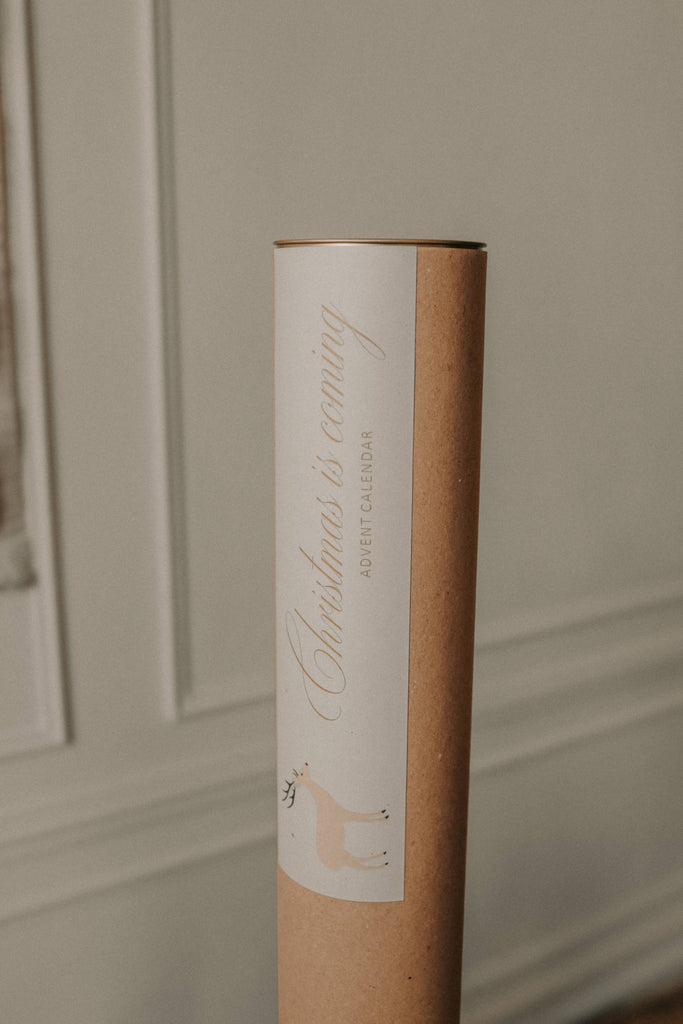 A brown cardboard tube with a white "Christmas is coming" label and a reindeer illustration, perfect for storing your Sabo Concept Christmas Cotton Advent Calendar, pictured against a softly lit, neutral-toned wall.