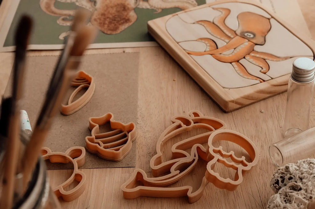 The Under The Sea Cutter Set - Eco Playdough Tool, made from biodegradable PLA, sits alongside brushes in a jar, a drawing of an octopus, and a wooden octopus carving on a wooden table—creating an arts and crafts scene with a playful marine theme.