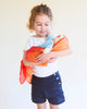 A young girl with curly hair, dressed in a white shirt and navy shorts, holds a doll wrapped in Sarah's Silks Enchanted Playsilk - Fire against a plain light background.