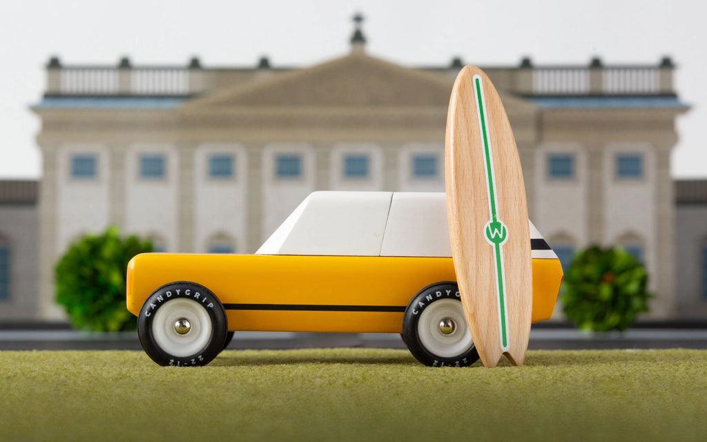 The Candylab Cotswold, a toy yellow car with white windows and black tires, is parked on green turf with a wooden surfboard standing upright next to it. In the background, a blurred image of a classical-style building flanked by trees completes the perfect scene for this charming toy vehicle.