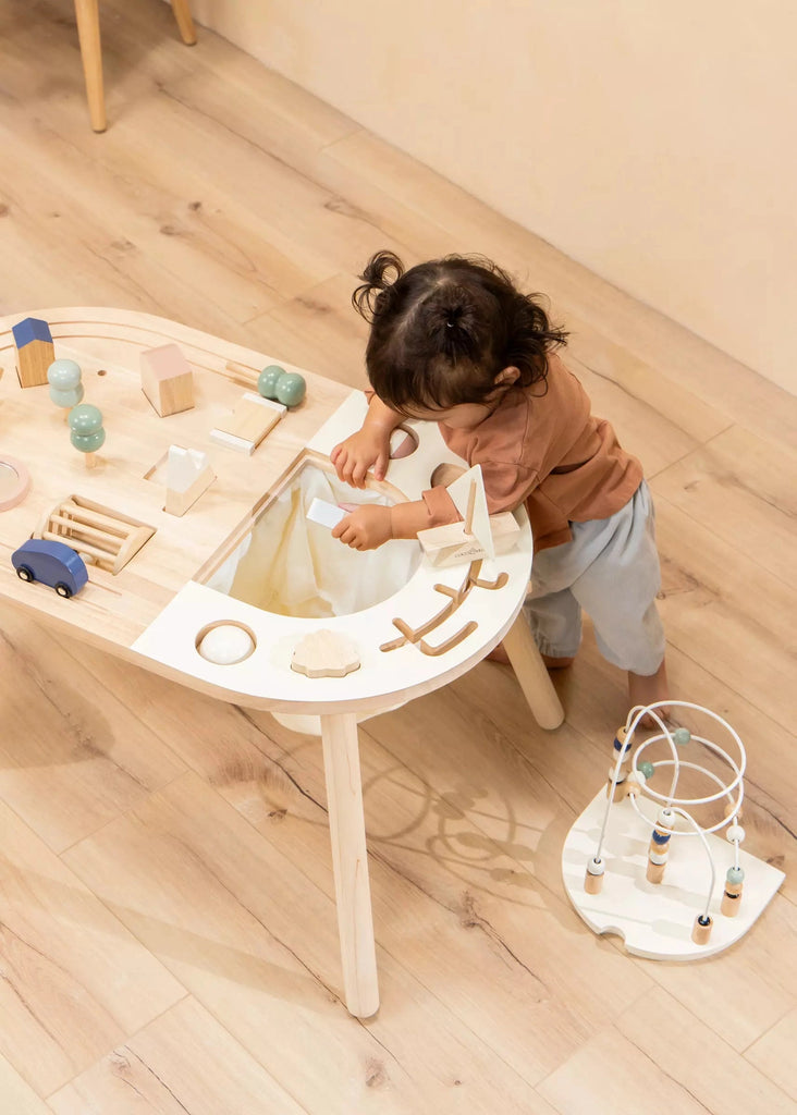 A toddler with dark hair plays with a Wooden Activity Table, including blocks and a bead maze, in a brightly lit room with a wooden floor.