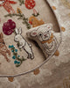 A close-up of the Coral & Tusk Sugar Egg Pocket Pillow shows beige embroidered fabric with a white rabbit, colorful flowers, daisy patterns, and a plush lamb—creating charming Easter decor with detailed stitching.
