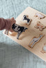 A child’s hand holds a cow-shaped piece from the Farm Animals Wooden Puzzle: A Tabitha Paige Puzzle above the matching animal puzzle board on a textured blanket, exploring this delightful toddler educational toy.
