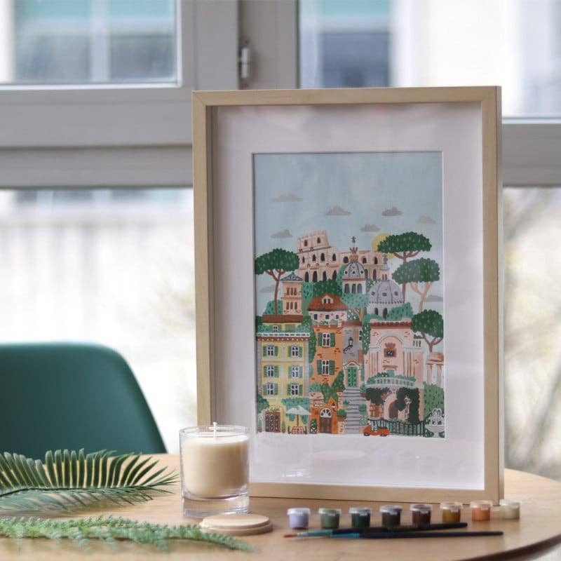 The Paint By Number Kit - Rome features a framed illustration of vibrant buildings and trees, displayed on a wooden table near a window with a white candle, green fern leaves, and paint pots with brushes—perfect inspiration for artists.