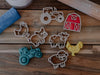 The Farm Cutter Set - Eco Playdough Tool features biodegradable cookie cutters shaped like a tractor, barn, chicken, pig, cow, and sheep arranged on a wooden surface alongside toy replicas of a tractor, chicken, and a blue item. This set is perfect for sensory play or as eco-friendly playdough tools to inspire creativity and imaginative fun.