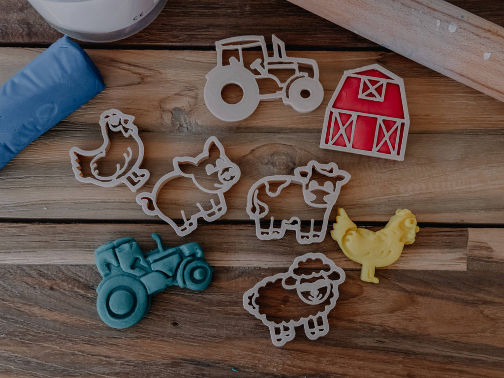 The Farm Cutter Set - Eco Playdough Tool features biodegradable cookie cutters shaped like a tractor, barn, chicken, pig, cow, and sheep arranged on a wooden surface alongside toy replicas of a tractor, chicken, and a blue item. This set is perfect for sensory play or as eco-friendly playdough tools to inspire creativity and imaginative fun.