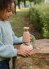 A young child with curly hair and a green sweater examines the Little Scientist Wooden Microscope on a large rock outdoors, surrounded by greenery and trees.