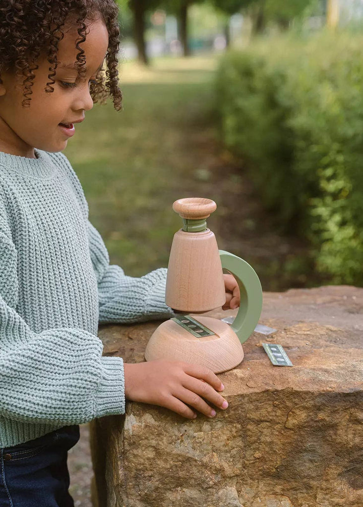 A young child with curly hair and a green sweater examines the Little Scientist Wooden Microscope on a large rock outdoors, surrounded by greenery and trees.