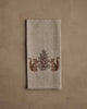 The Coral & Tusk Chipmunks with Pinecone Tea Towel features embroidered chipmunks with a pinecone and leafy motif, accented by metallic silver stitching on beige linen, displayed on a neutral textured background.