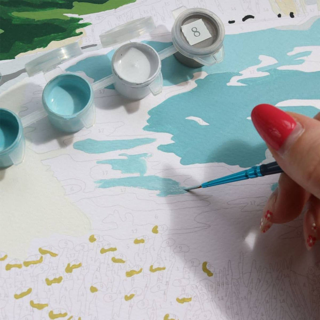 A hand with red nails paints the "Paint By Number Kit - Picnic in Étretat by Ananya Bhandari," featuring blue and white Normandy coast scenes. Open paint pots surround the canvas, showing numbered sections ready to be filled.