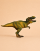 The Schleich Tyrannosaurus Rex Dinosaur Toy, featuring green and yellow coloring and resembling a predatory T. rex, stands upright against a plain beige background with its mouth open to display its teeth.