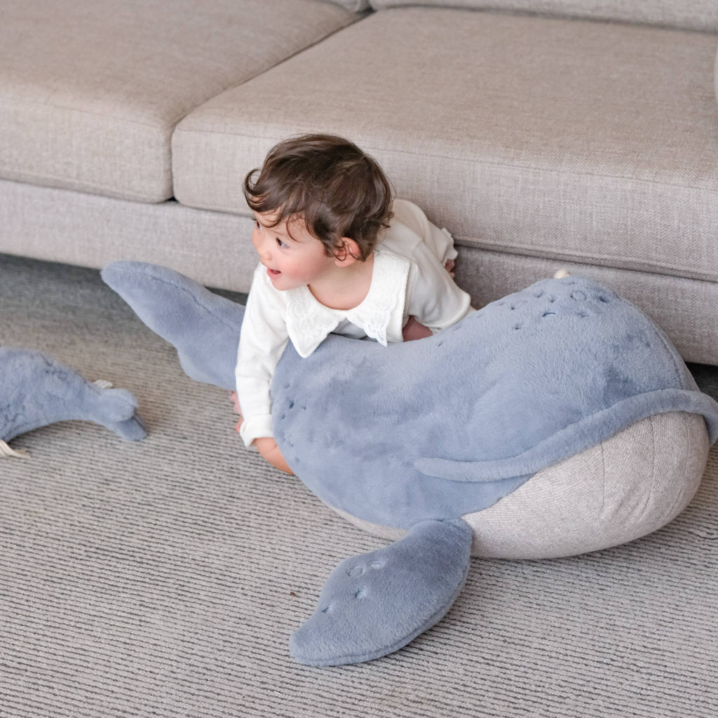 A young child in white clothes lies on the Ride-On Whale Pouf, a large plush toy with a playful whale design, placed on a carpet next to a light-colored sofa.