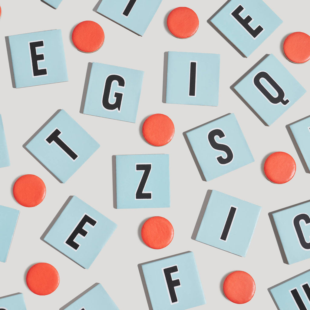 Scattered light blue square letter tiles and orange circular tokens from a Scrabble for Juniors Vintage Bookshelf Edition on a light background.