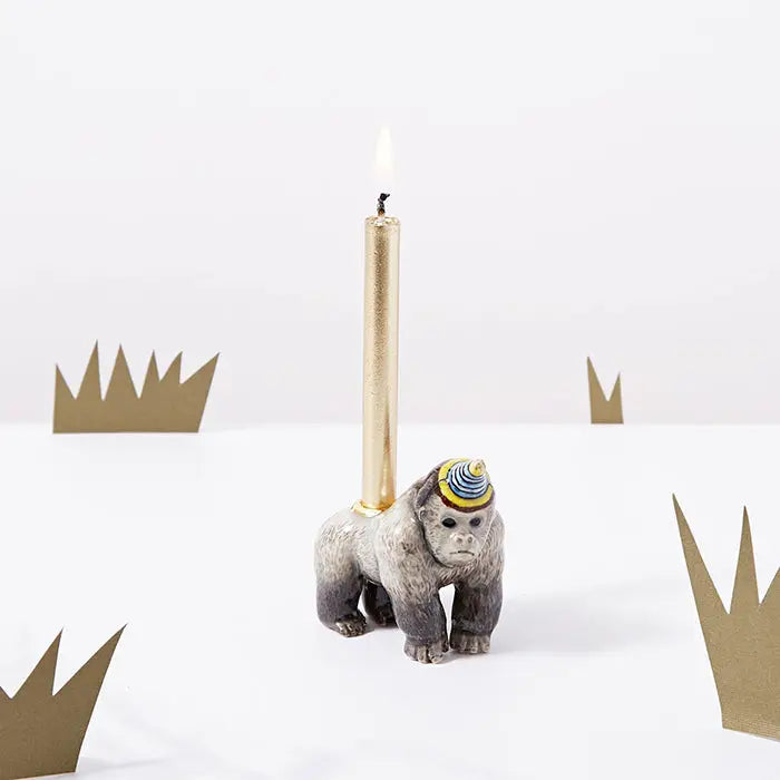 A hand-painted, Gorilla "Celebrate Nature" cake topper, crafted in fine porcelain and painted in gray and gold, holds a lit, slender white candle. Golden cut-out crowns are in the background.