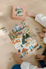 Two children assemble the Secrets of the Autumn Woods 300 Piece Puzzle, featuring a lively outdoor scene with people, trees, and animals on a wooden floor. The puzzle box and large reference poster are placed nearby.