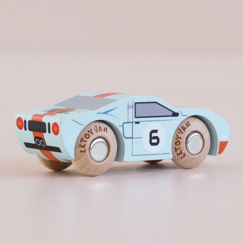 A light blue classic toy car, the No. 26 GT145 Fred made in England, features the number 26 on the door, wooden wheels, and orange racing stripes on the back—all capturing the spirit of an American performance car against a plain background.