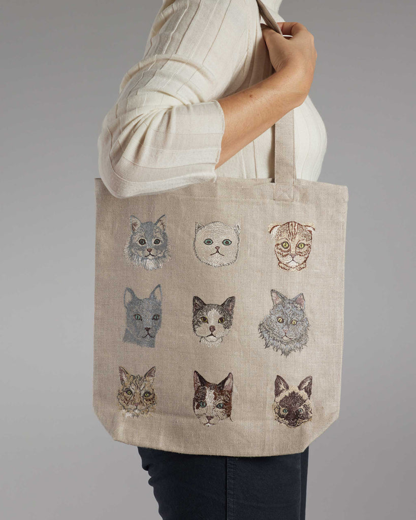 A person in a light shirt holds a beige Cats Tote Bag with nine uniquely embroidered feline faces. Displayed from the side with a bent arm, they proudly show off the bag's charming design and handy inside pocket.