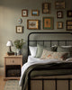 Cozy bedroom with a metal bedframe, green and white bedding, a Coral & Tusk Mushrooms and Ferns Lumbar Pillow, a wooden nightstand with books, lamp, and flowers. Framed art forms a gallery wall above the bed.