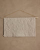 The Coral & Tusk North Pole Advent Calendar features a plain, light beige fabric hanging on a wooden dowel with a thin string, displayed against a textured wall for a minimalist and subtle Christmas countdown. The fabric is wrinkled and unadorned.
