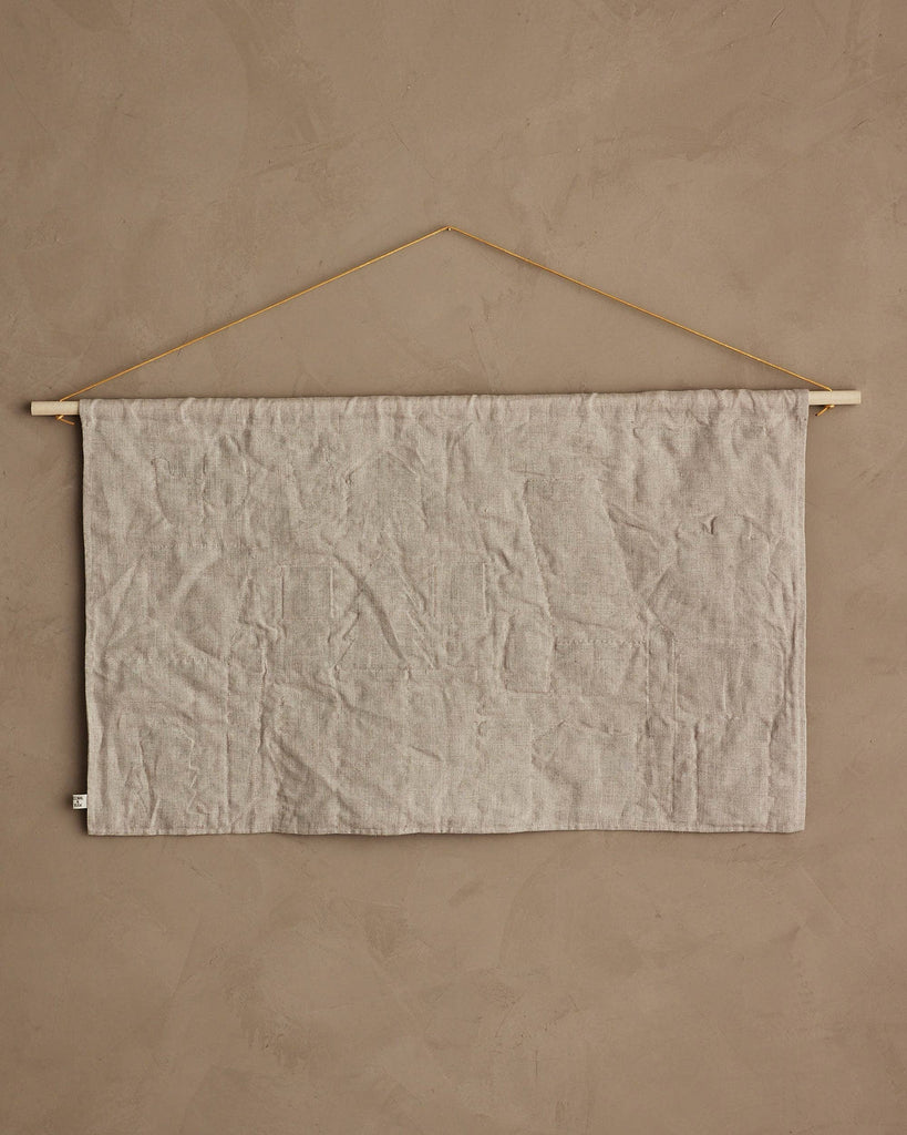 The Coral & Tusk North Pole Advent Calendar features a plain, light beige fabric hanging on a wooden dowel with a thin string, displayed against a textured wall for a minimalist and subtle Christmas countdown. The fabric is wrinkled and unadorned.