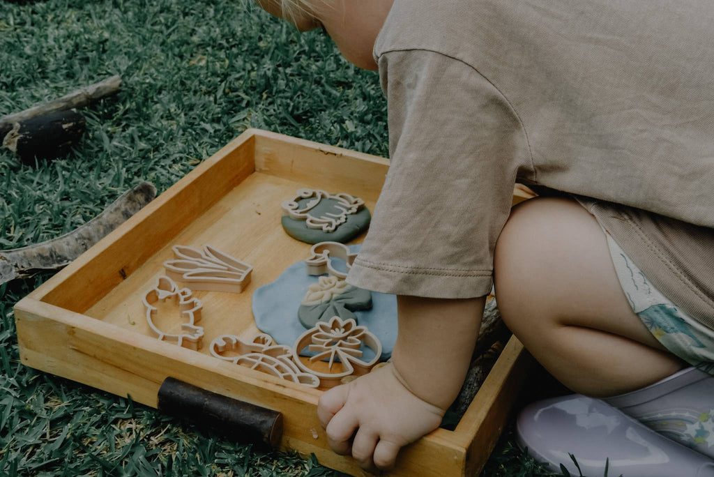 A child kneels on grass, playing with the Pond Life Cutter Set – Eco Playdough Tool and decorated cookies in a wooden tray.