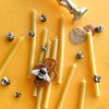 The Beeswax Birthday Candle Set, featuring thin yellow eco-friendly candles and small decorative bees, is displayed on a yellow surface with a honeycomb ornament, brass candle holder, and textured wax details.
