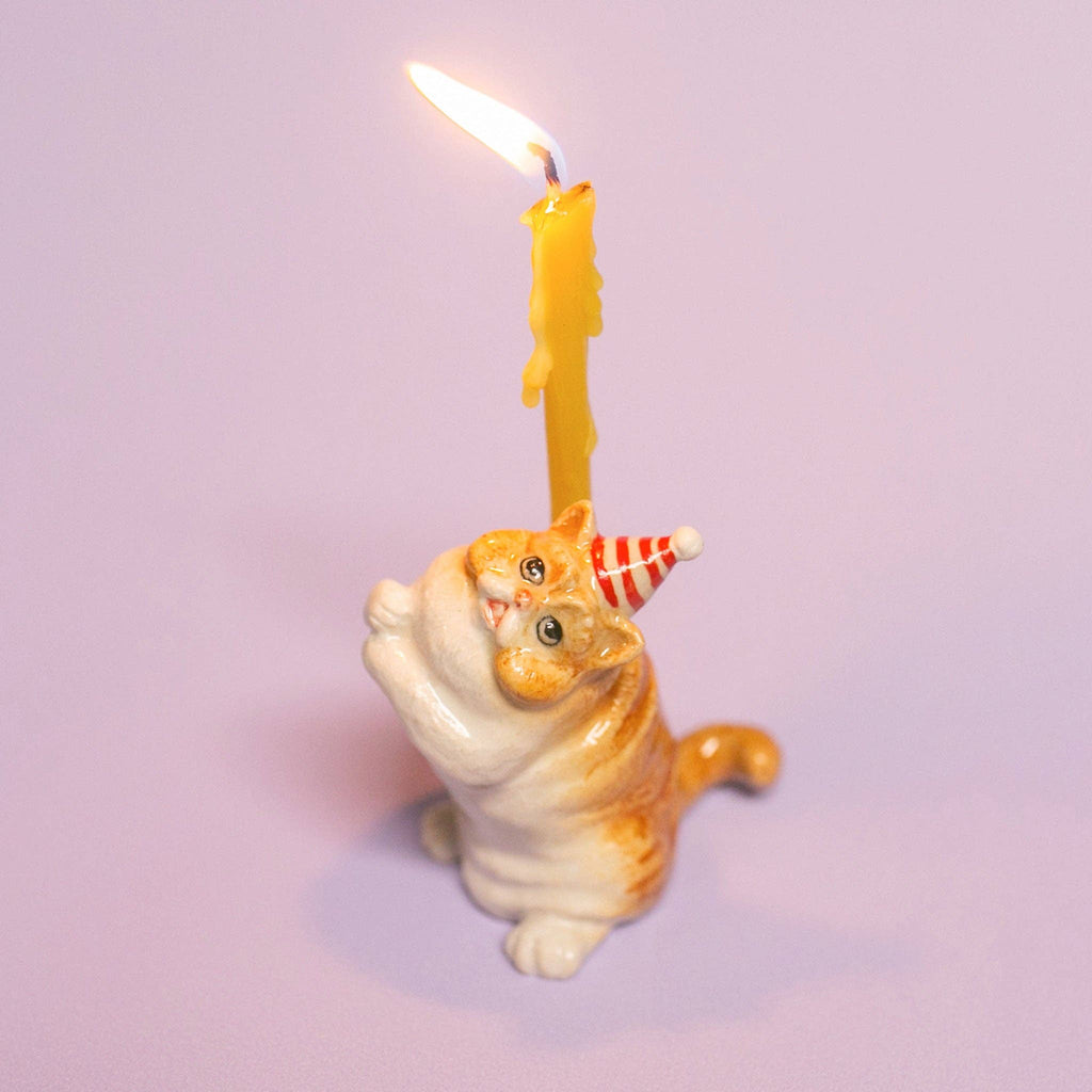 The Exotic Shorthair Cat Cake Topper is a ceramic orange and white figurine with a red party hat, holding a lit yellow candle—ideal for cat-themed parties and cake decoration, shown on a light purple background.