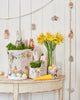 A festive Easter setup on a wooden table includes the Coral & Tusk Easter Parade Basket, embroidered with bunny and animal designs. It's filled with greenery and plush toys, complemented by a vase of yellow flowers, pastel eggs, and hanging decorations against a white wall.