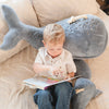 A young boy sits on a bed, reading a book while leaning against a large Ride-On Whale Pouf. He looks focused on the story, and the bedding has a light patterned design.