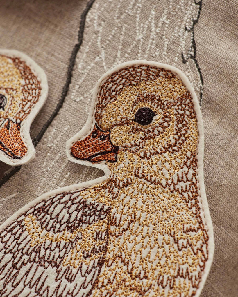 Close-up of a fabric patch on the Coral & Tusk Mama Duck Pocket Pillow, showcasing a finely embroidered duckling with brown and beige stitching, set against an embroidered outline of a larger duck.