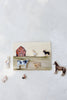 A rectangular cookie, inspired by the “Farm Animals Wooden Puzzle: A Tabitha Paige Puzzle,” is decorated with a cheerful red barn and an array of farm animals—a cow, dog, goose, and pig—resembling an engaging farm animal puzzle. Encircling it are small cookies shaped like a horse, cat, llama, chick, and goat atop a light background, evoking the playful spirit of this inviting toddler educational toy.