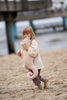 A young child in a light shirt and purple pants stands barefoot on the sand, hugging the Senger Naturwelt Cuddly Animal - Seahorse. In the background, water and a blurred structure can be seen.
