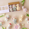 The Cuddle + Kind Baby Chicken, an heirloom-quality, hand-knit doll handmade in Peru, sits among other pastel-colored baby animal dolls on a beige surface with white tulips. Four are arranged in a pink box while others surround it.