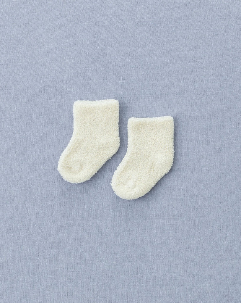 A pair of small, white baby socks are laid out side by side on a light blue fabric background. These fluffy socks are soft and appear new and unworn, perfect as part of Lithuania baby accessories.