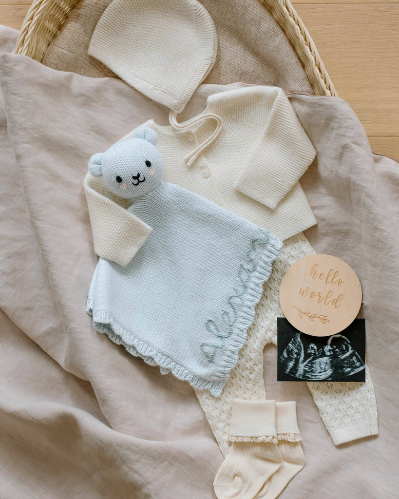 A cream-colored baby outfit with bonnet, top, pants, socks, a Cuddle + Kind Organic Cotton Baby Bear Lovey (Baby Blue) personalized with "Alexander," a "hello world" wooden plaque, and an ultrasound photo—perfect for a charming fair trade baby gift.