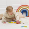 A young child sits on a soft white rug, playing with the Counting Wooden Numbers Shape Sorter. Behind them are beige pillows and a rainbow cushion against a white wall, creating an inviting early learning space.