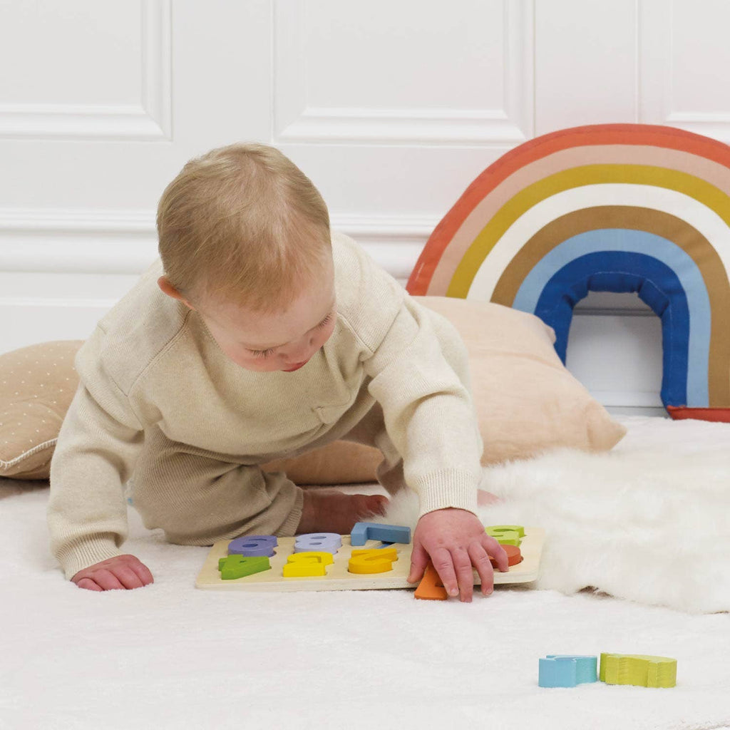 A young child sits on a soft white rug, playing with the Counting Wooden Numbers Shape Sorter. Behind them are beige pillows and a rainbow cushion against a white wall, creating an inviting early learning space.