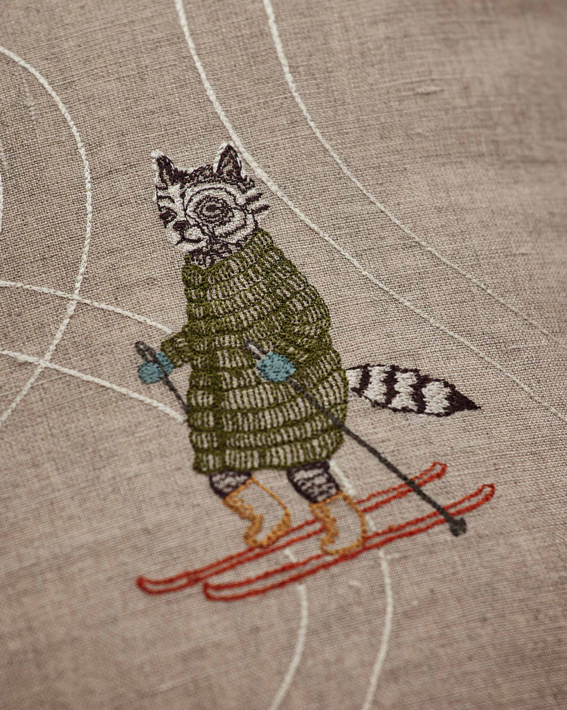 The Coral & Tusk Downhill Skiers Pillow features a raccoon in a green coat, blue mittens, and brown boots skiing on red skis with white trails—an ideal alpine accent for any skier’s decor.